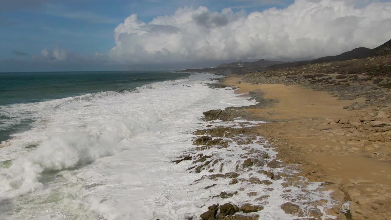 Punta Gordo coastal frame with dense white foam covering much of the near shore, exposed rocks cutting through the surf, tan sand to the right, and no visible people.