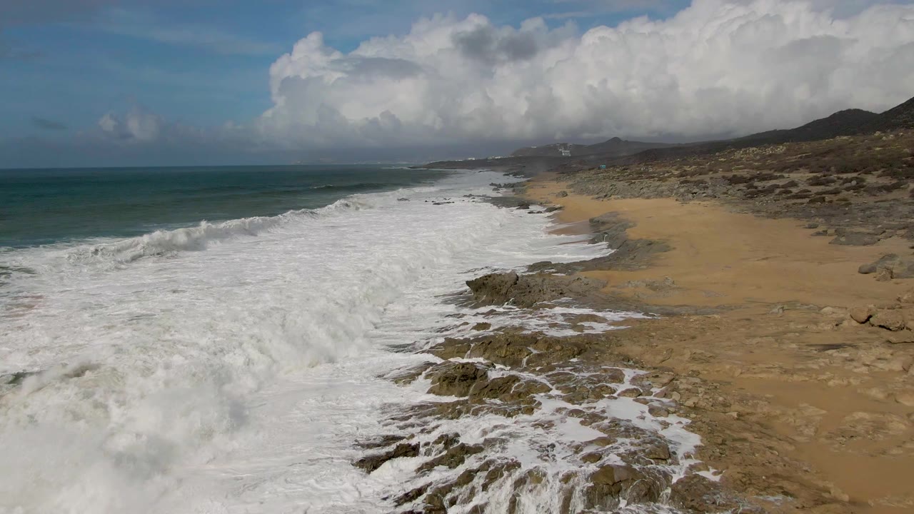 Aerial Punta Gordo beach view with the breaking wave lower in frame, white foam wrapping around dark rocks, sand extending inland, and no visible people.