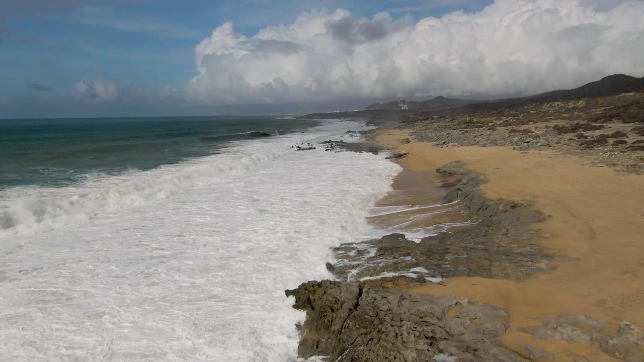 Aerial frame over Punta Gorda, Baja California Sur, showing broad white surf entering from the left, tan sand and dark rocks along the shore, distant hills, clouds, and no visible people.