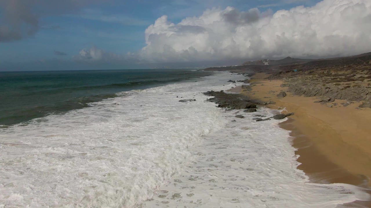 Aerial view of Punta Gorda East Cape surf spreading across a sandy shoreline with dark rocks, green-blue ocean to the left, low hills in the distance, and no visible people.