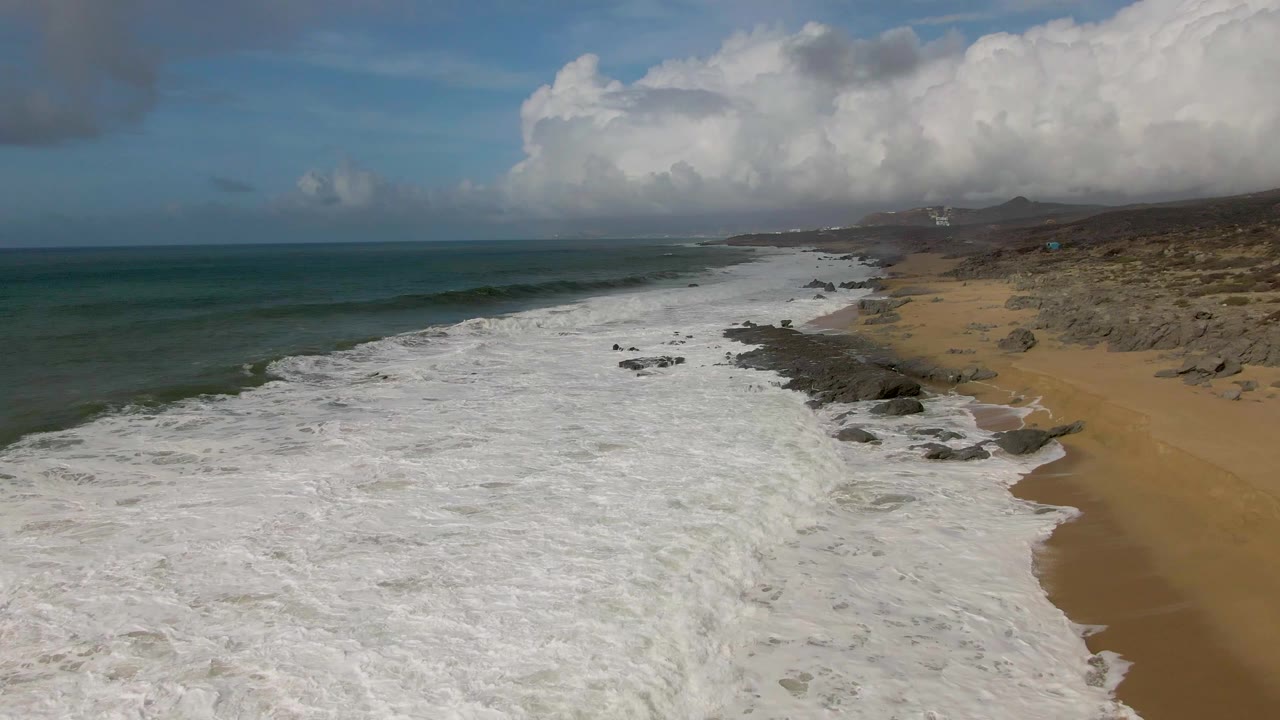 Aerial frame over Punta Gorda East Cape showing dense white foam across the lower beach, dark rock clusters near the waterline, tan sand to the right, and no visible people.