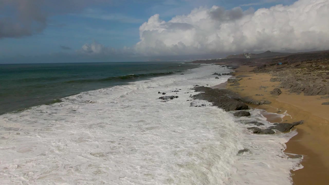 Punta Gorda East Cape coastline from above, with surf breaking across the center, a darker rock band along the beach, open ocean on the left, cloud banks above the horizon, and no visible people.