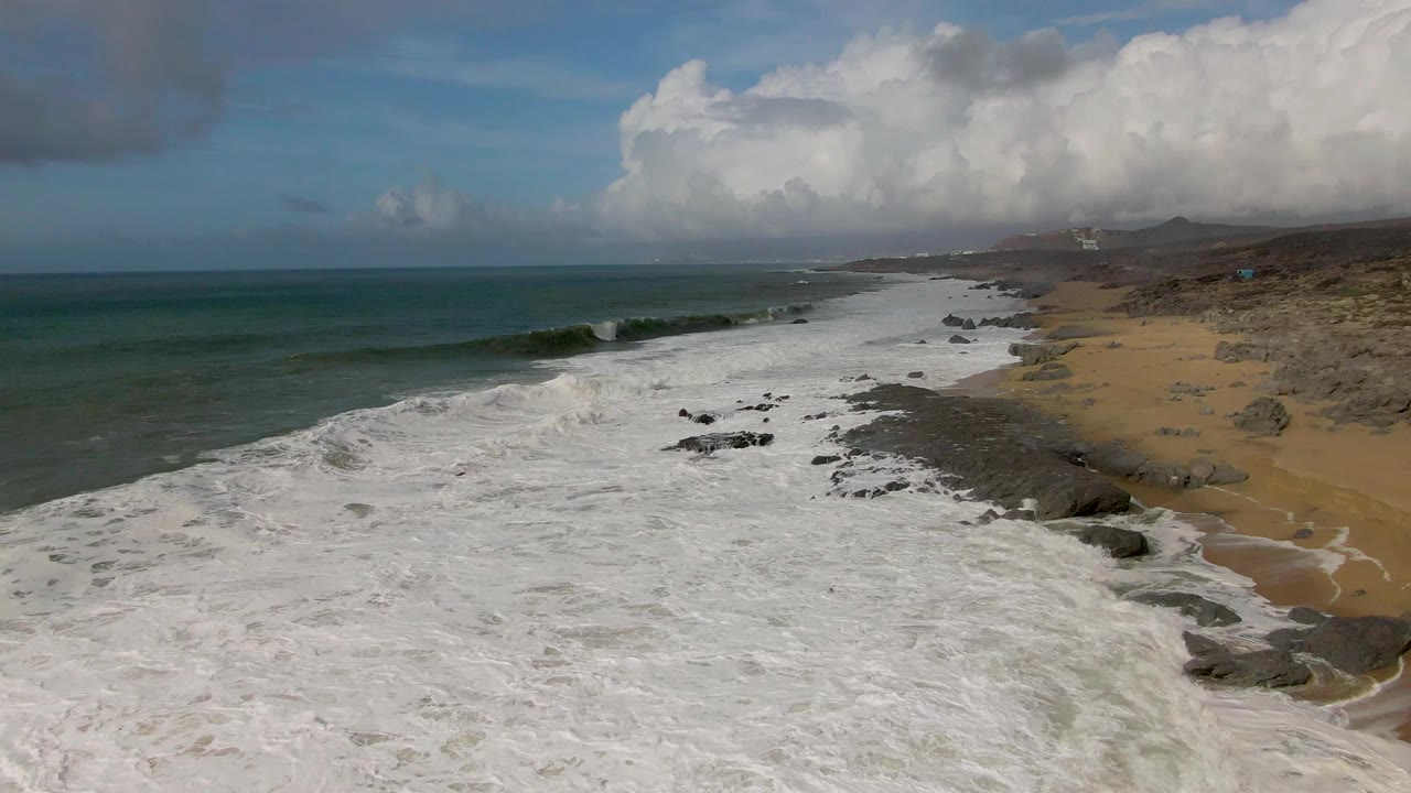 Long sandy and rocky shoreline with broad white surf under a blue cloudy sky.