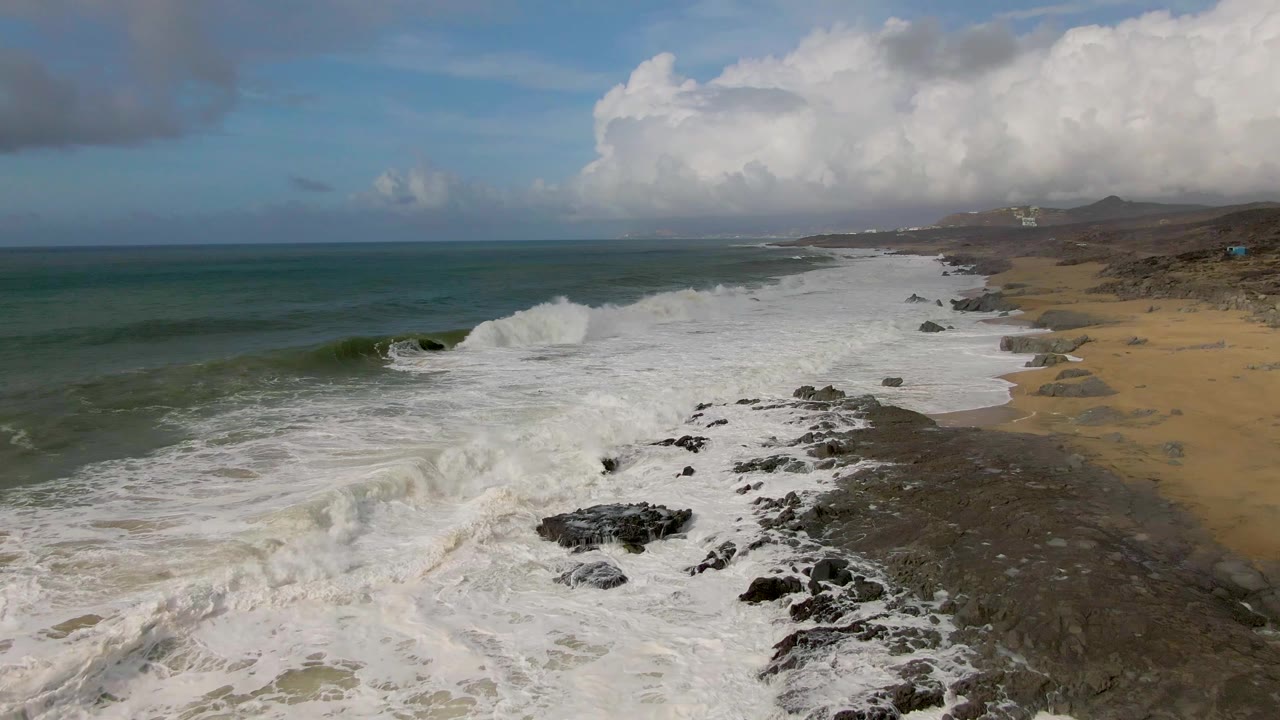 Aerial view at Punta Gorda, Baja California Sur, with white waves breaking over dark shoreline rocks beside tan sand and no visible people.