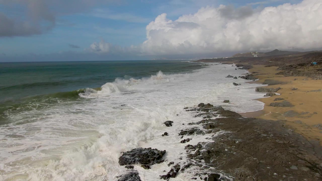 A large wave crests near dark rocks at Punta Gorda, with white foam spreading toward the sandy beach and no visible people.