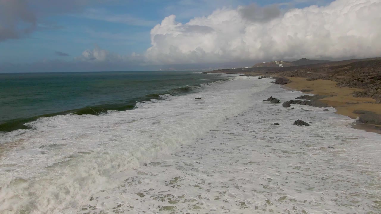 Aerial view of green-blue ocean waves breaking onto a sandy rocky shoreline.