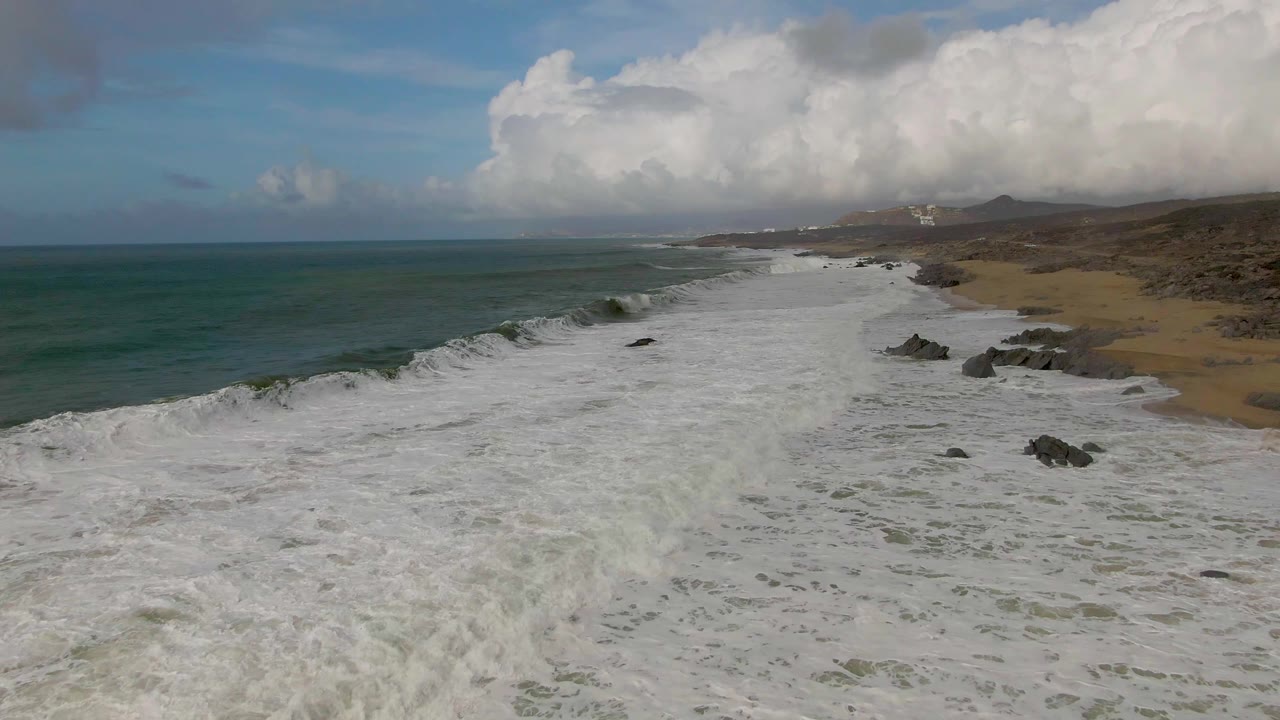At Punta Gorda, Baja California Sur, white surf advances across the sandy beach while dark rocks break up the shoreline, with open ocean to the left and no visible people.