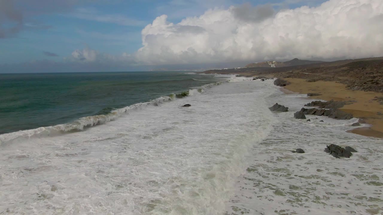 A Punta Gorda aerial frame shows a larger wave crest approaching the beach, white foam spread across the lower frame, rocky outcrops to the right, and no visible people.