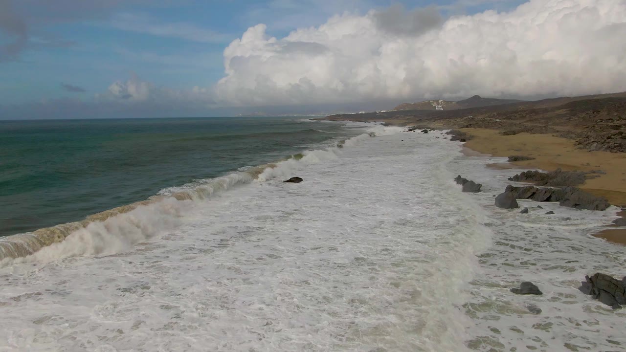 At Punta Gorda, Baja California Sur, a wide sheet of whitewater covers the shore, green-blue water remains visible offshore, dark rocks sit along the right side, and no visible people appear.