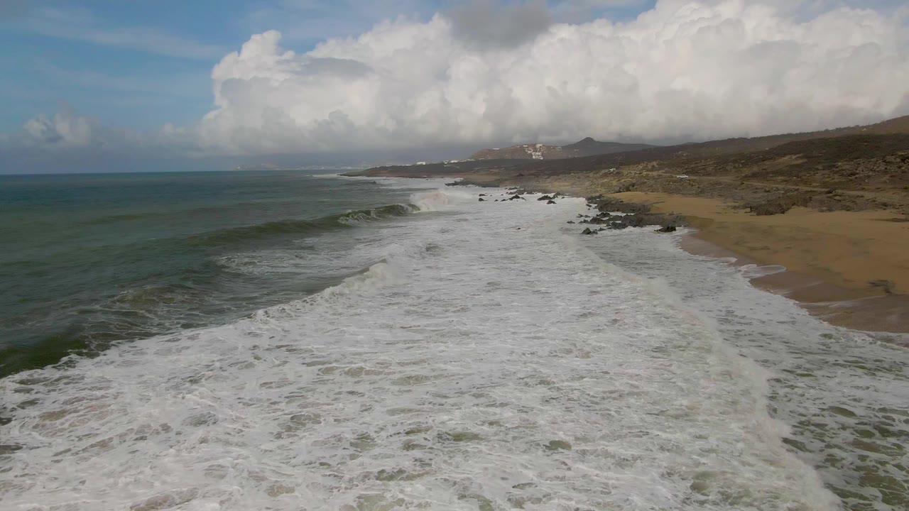 At 0:00, an aerial view looks along the Punta Gorda East Cape shoreline as white surf spreads across the foreground, green-blue ocean sits left, tan sand and rocks line the right side, and no visible people appear.
