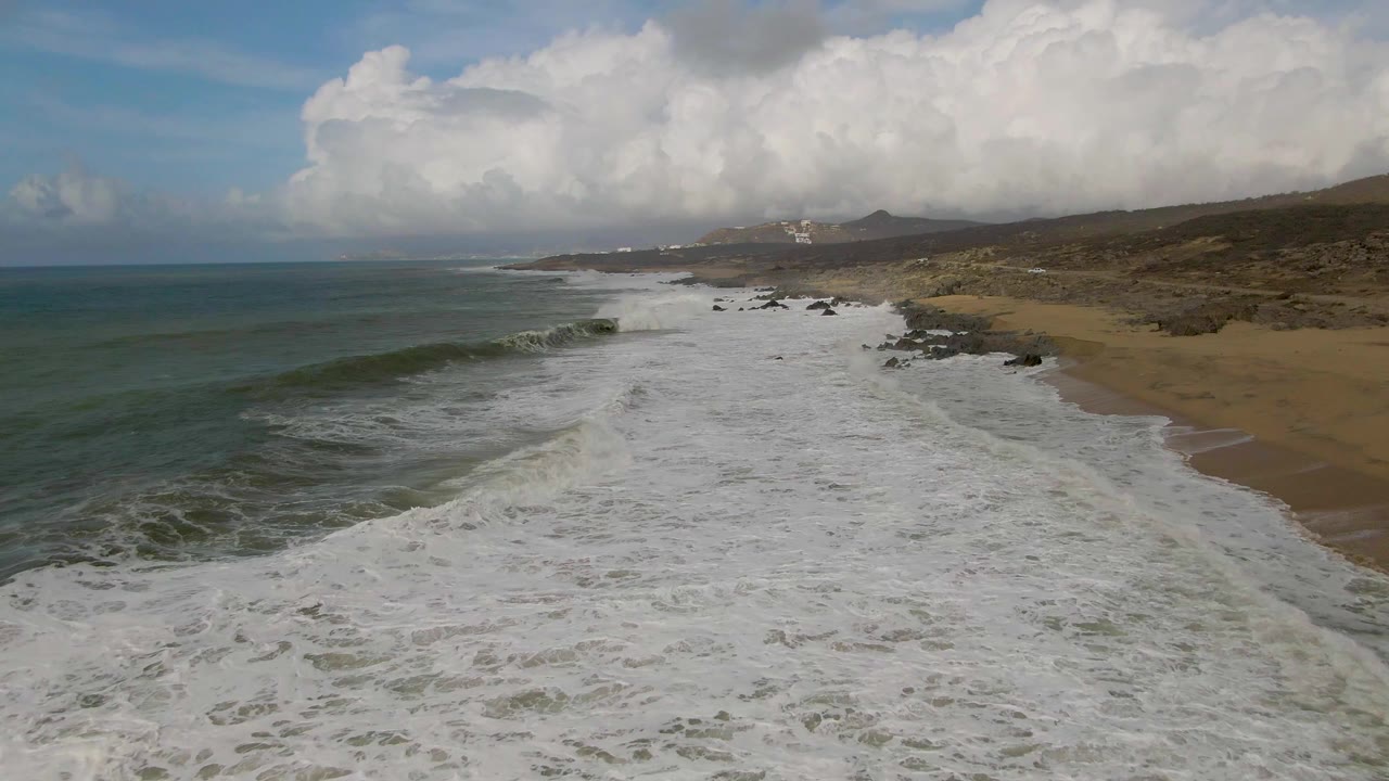 At 0:01, the Punta Gorda East Cape coast remains in a high oblique view as a broad white band of breaking water pushes shoreward beside dark rocks and tan beach, with no visible people.