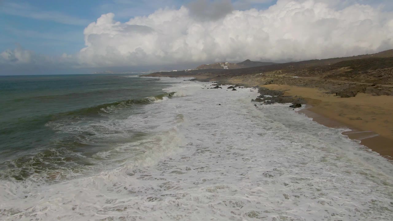 At 0:02, white foam covers most of the lower frame at Punta Gorda East Cape while a darker green wave face forms on the left and the rocky sandy shore continues into distant hills, with no visible people.