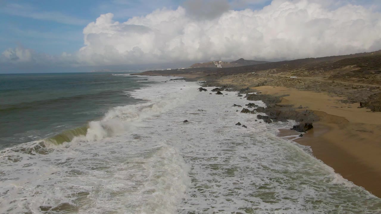 Aerial view at Punta Gordo, Baja California Sur, with blue-green ocean on the left, white surf rolling over rocks and sand, low hills in the distance, and no visible people.