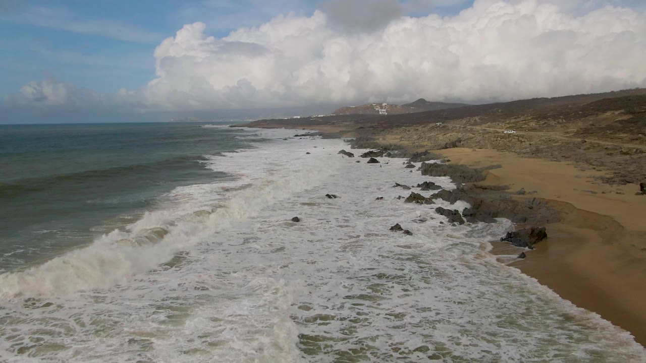 Whitewater spreads along a rocky shoreline with open ocean and cloudy sky.