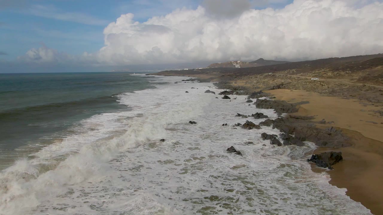 Punta Gordo aerial shoreline view with dense foam in the center, scattered black rocks near the beach, blue-green water to the left, distant hills, and no visible people.