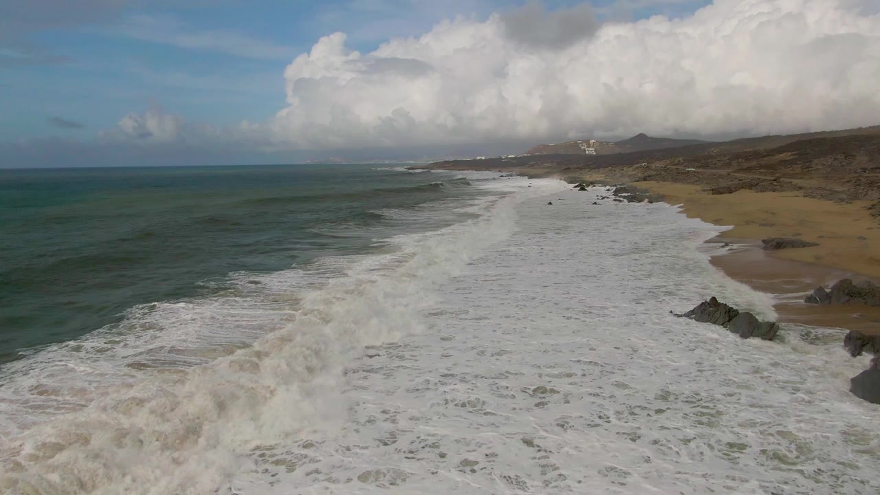Aerial view of Punta Gorda, Baja California Sur coastline with green-blue ocean on the left, white foam covering the shore, sandy beach and rocks on the right, and no visible people.