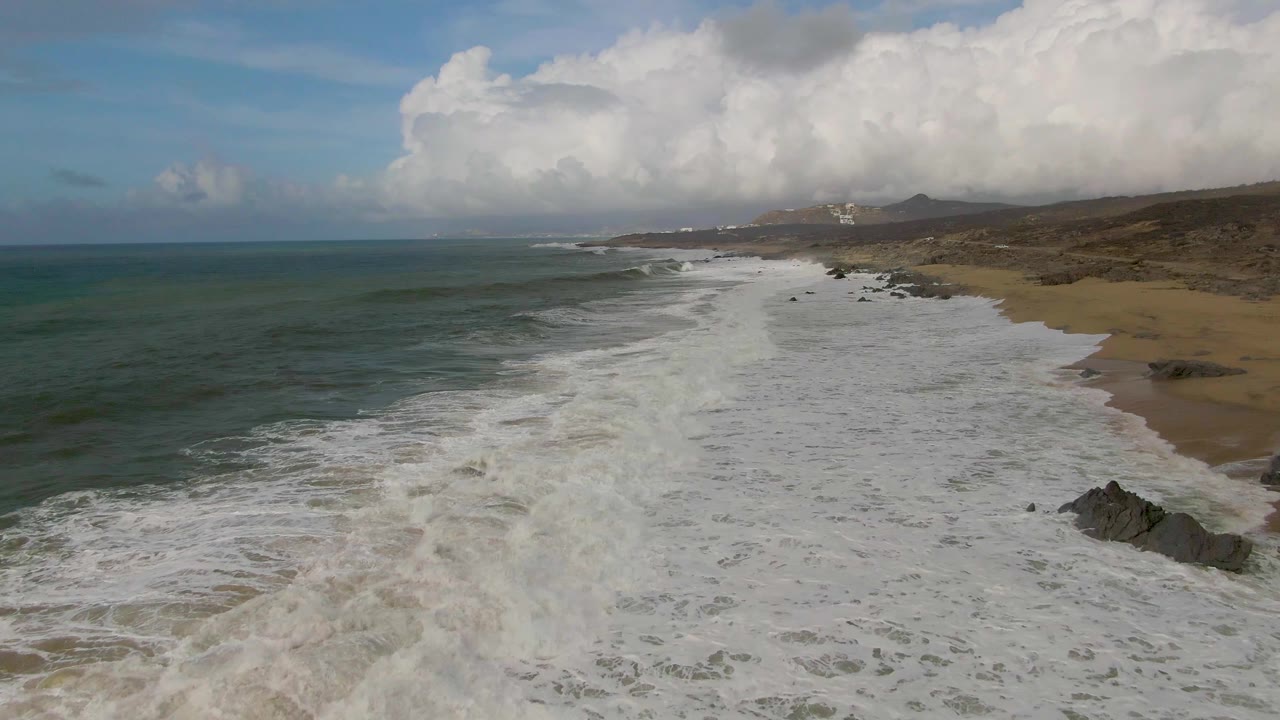 Aerial Punta Gorda beach view with a broad band of white surf moving up the sand, dark rocks near the lower right edge, cloud cover over the distant coast, and no visible people.