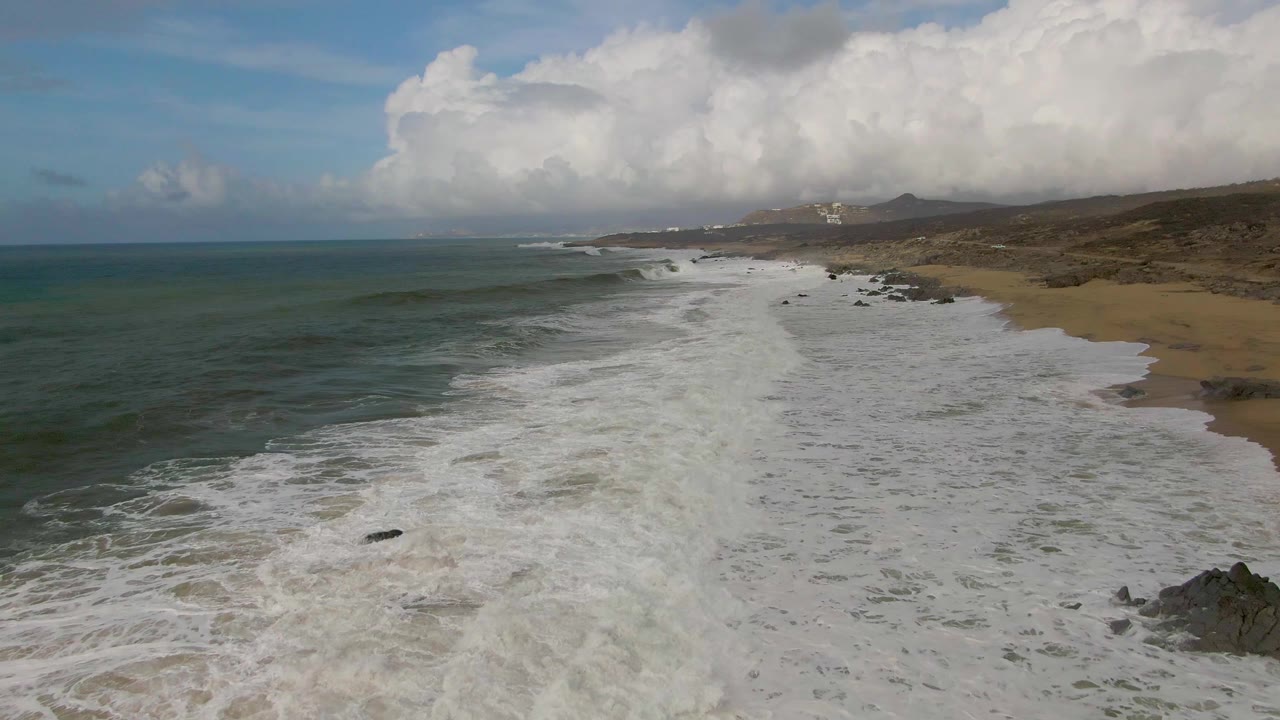 Aerial frame from Punta Gorda with the surf line stretching into the distance, blue-green ocean left of frame, sandy beach and scattered rocks to the right, and no visible people.