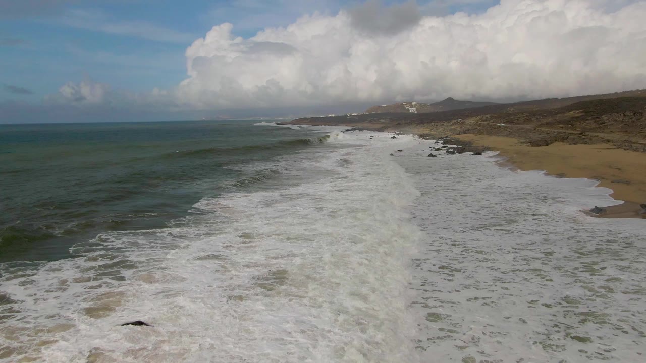 Aerial shoreline with ocean, white foam, sand, rocks, and a large cloud bank overhead.