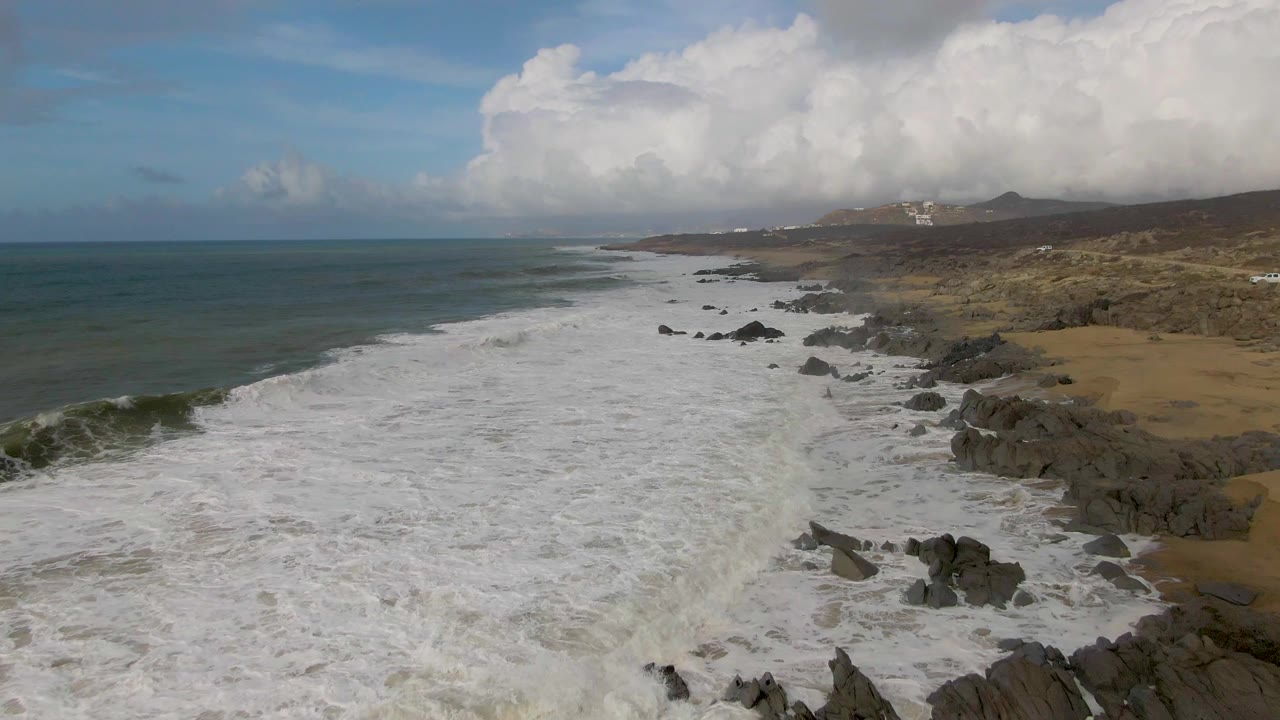 Aerial view at Punta Gorda, Baja California Sur, with white surf spreading over rocks and sand, teal ocean to the left, clouded sky above, and no visible people.