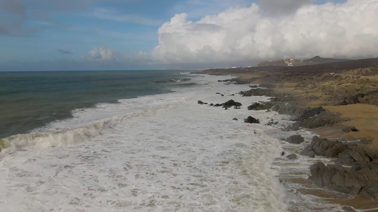At Punta Gorda, the foamy surf covers more of the rocky shoreline, with scattered black rocks in the water, brown land beyond, large clouds overhead, and no visible people.