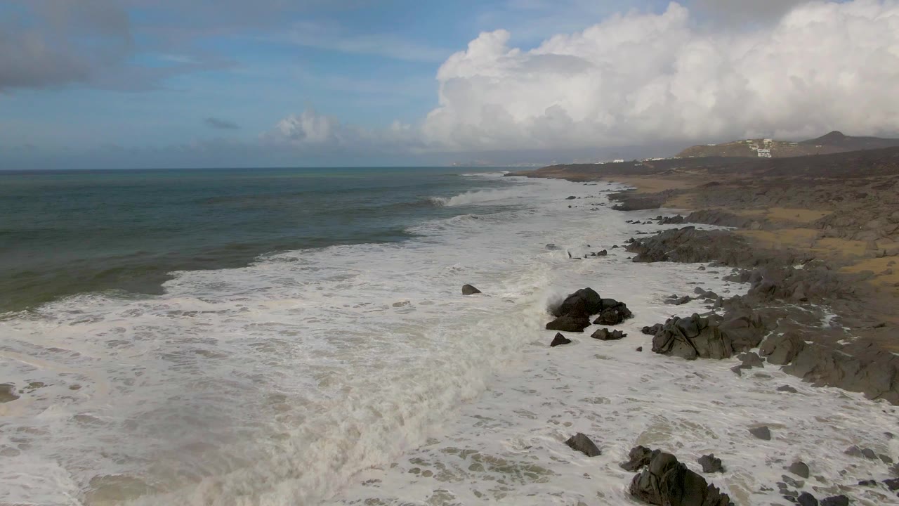 Aerial view of waves spreading white foam across dark rocks on the Punta Gorda East Cape shoreline in Baja California Sur, with no visible people.