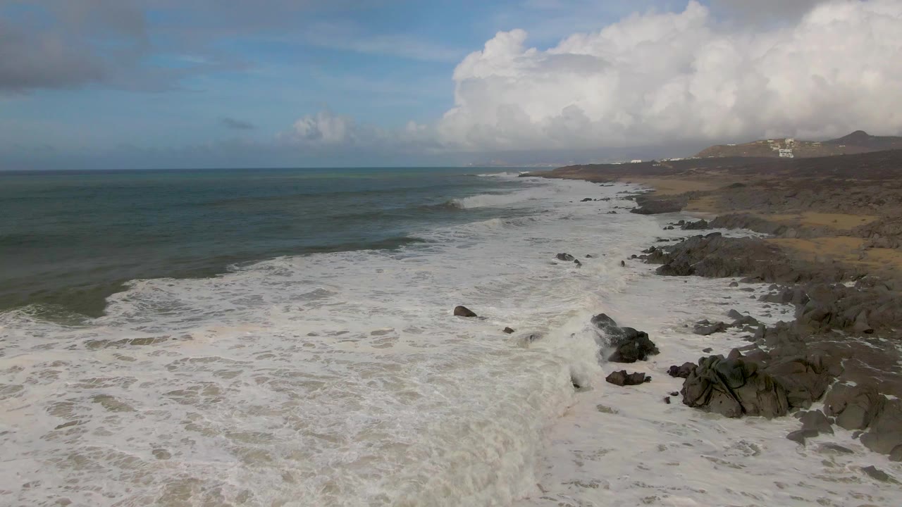 Wide elevated frame of Punta Gorda shoreline where whitewater pushes farther across the rocks, with open ocean to the left and no visible people.