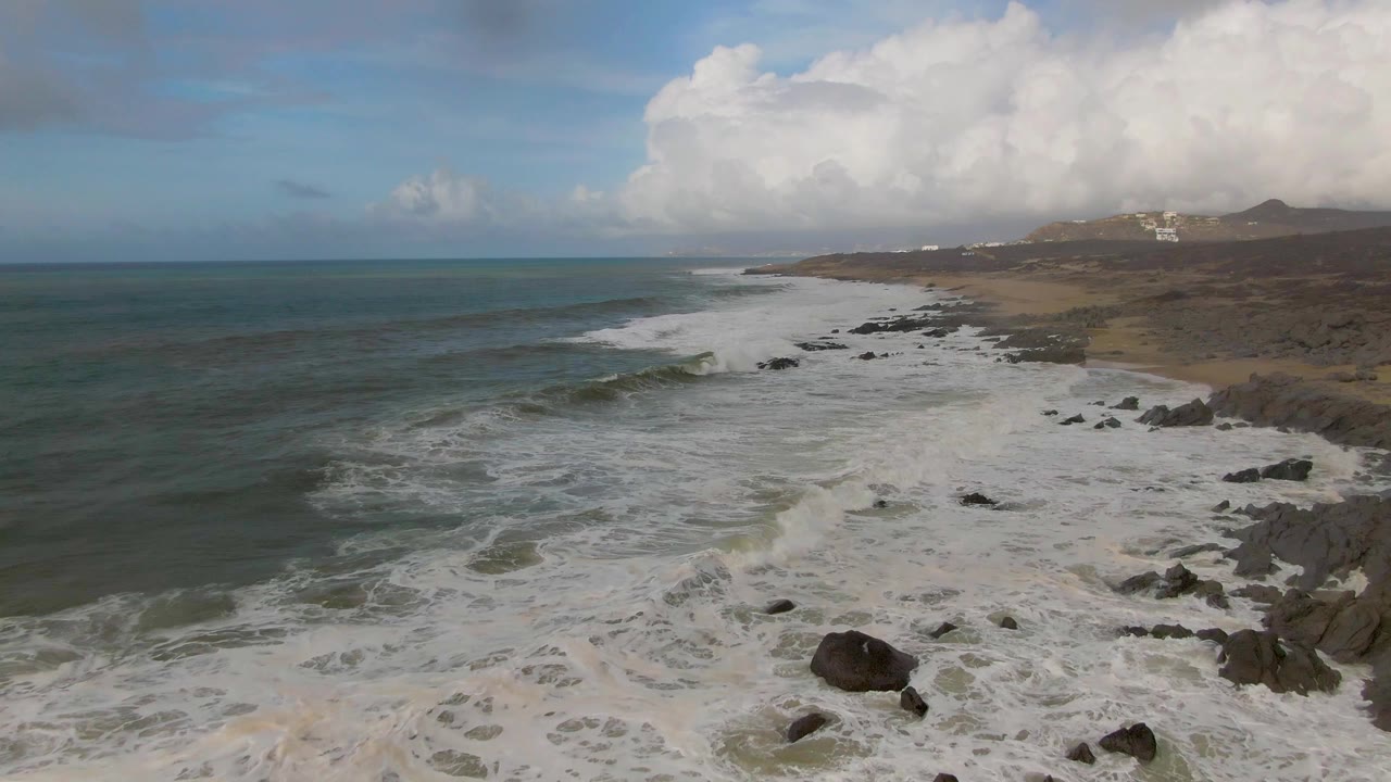 At 0:01, the elevated Punta Gorda East Cape view shows a new wave line curling near shore while white foam covers rocks and sand, with no visible people.