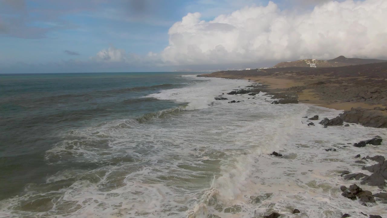 At 0:02, whitewater expands across the Punta Gorda East Cape rocks, the open sea fills the left side, and the clouded hills remain in the background with no visible people.