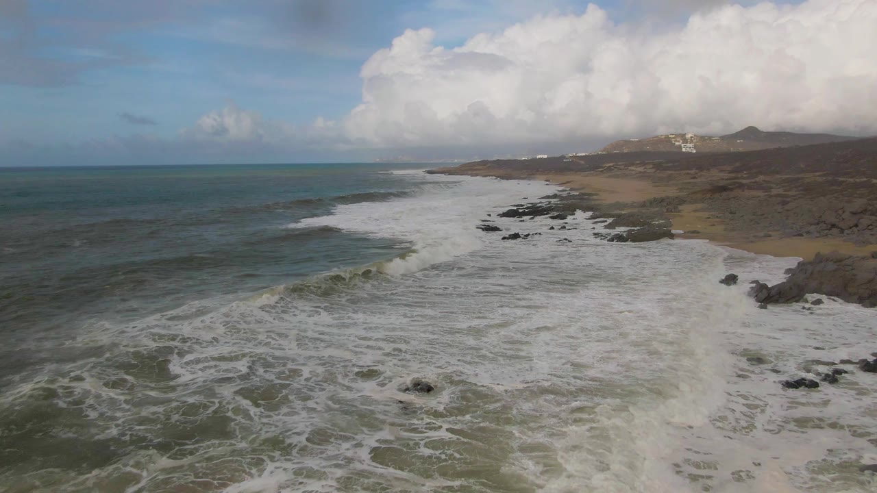At 0:03, the aerial composition looks along Punta Gorda East Cape as surf drains around scattered rocks, with ocean on the left, beach and hills to the right, and no visible people.
