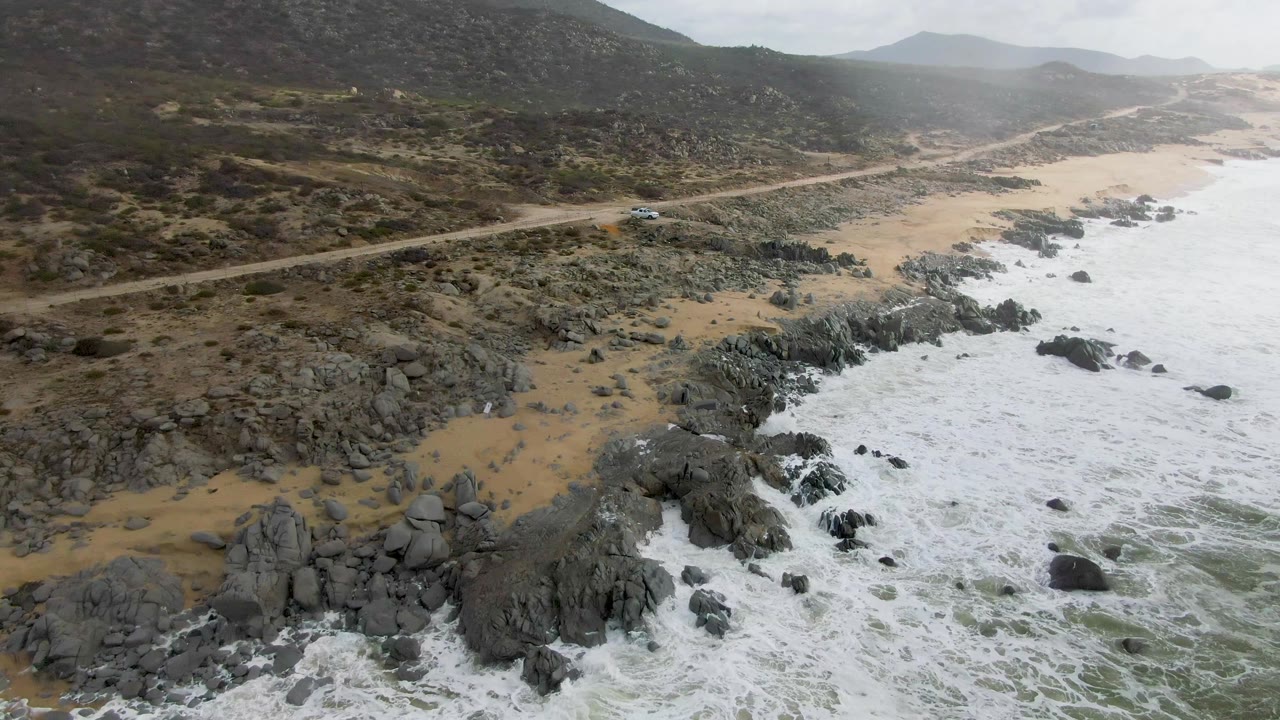 Aerial frame from Punta Gorda, Baja California Sur, showing a small car on a dirt coastal road above dark rocks, sandy beach, foamy surf, scrub hills, and no visible people.