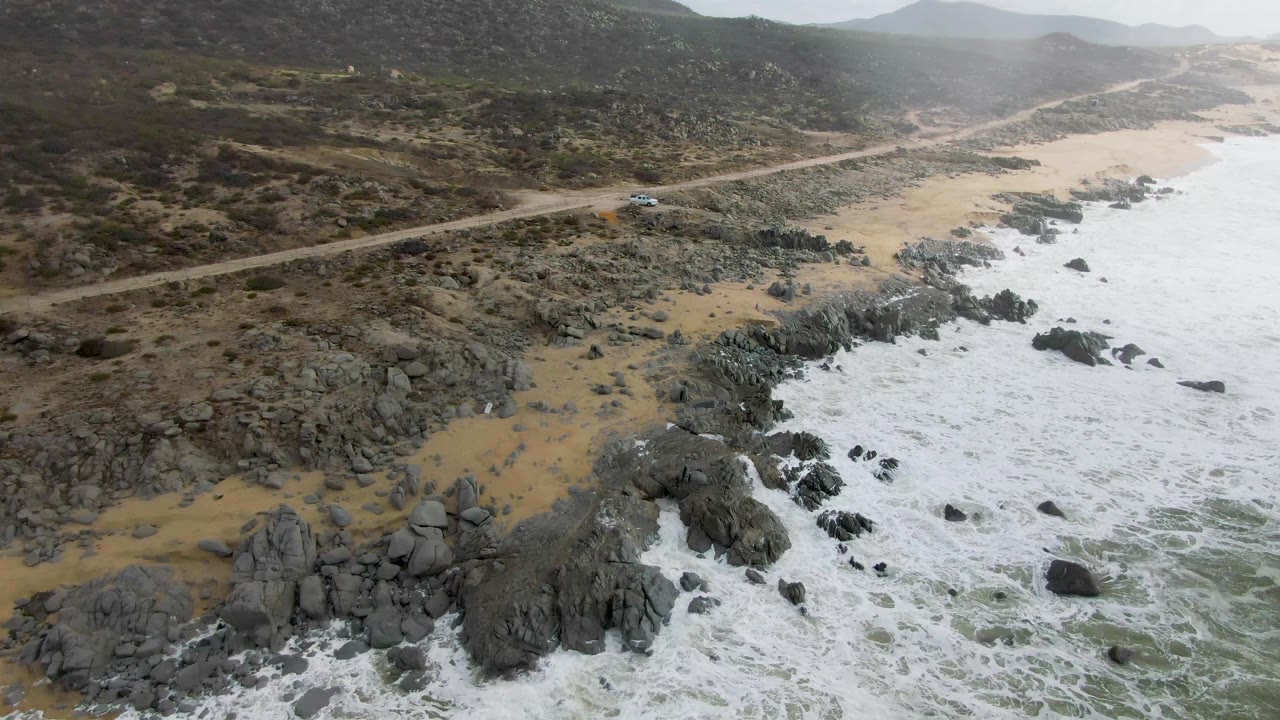 Aerial frame from Punta Gorda, Baja California Sur, with the car farther along the pale road while white surf spreads around black rocks below and no visible people appear.