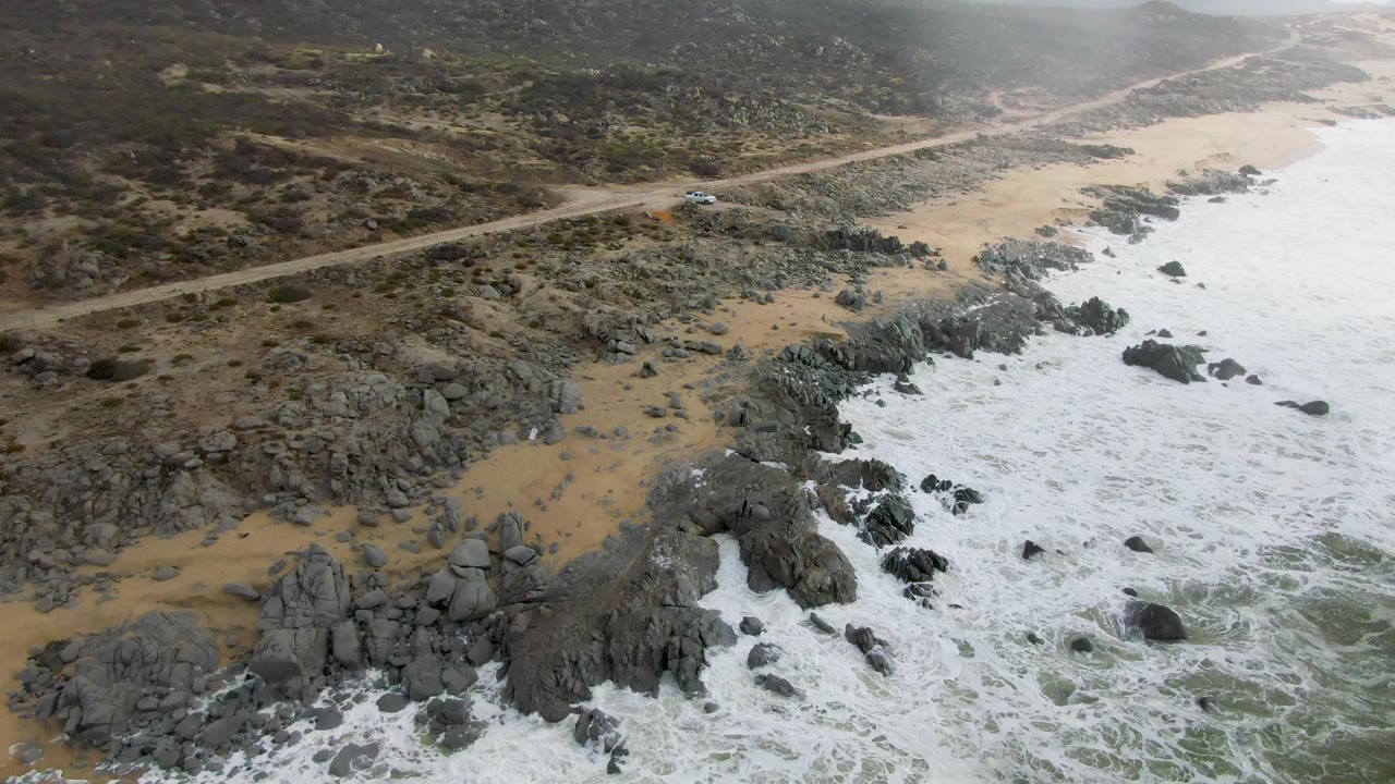 Aerial frame from Punta Gorda, Baja California Sur, showing the car near the upper road, a diagonal band of beach and rocks, heavy white foam along the waterline, and no visible people.