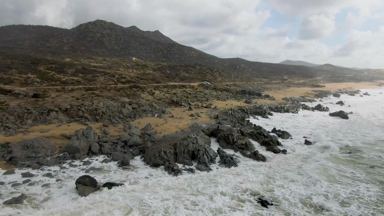 Aerial frame from Punta Gorda, Baja California Sur showing white surf around dark shoreline rocks, tan sand, a coastal road, low mountains, cloudy sky, and no visible people.