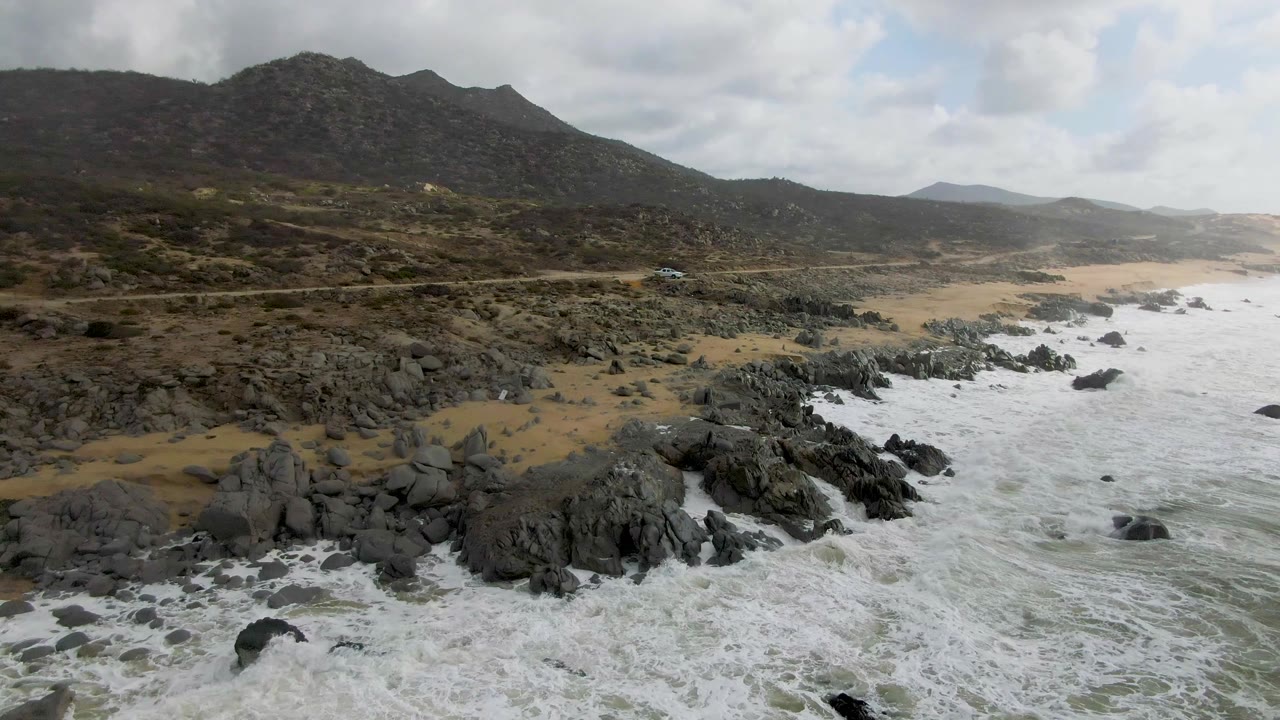 Aerial view in Punta Gorda, Baja California Sur with surf spreading farther across the foreground rocks, a pale sand strip, road, mountain slope, overcast sky, and no visible people.