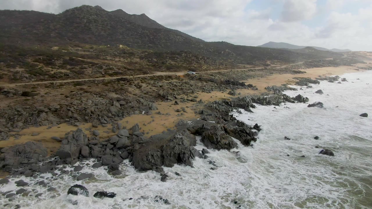 Punta Gorda, Baja California Sur coastline from above, with churning white water on the right, jagged black rocks near the beach, a road through scrub terrain, distant hills, and no visible people.