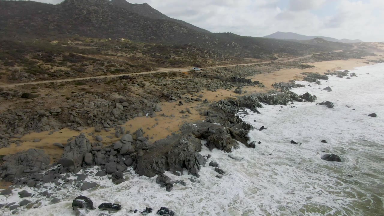 Rocky beach and surf below a road with a small vehicle on the coast.
