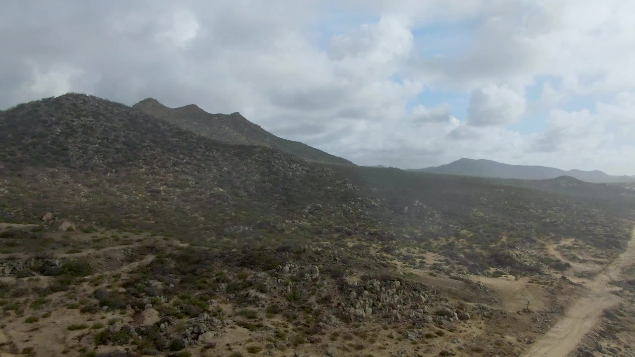 Aerial frame over Punta Gorda terrain with a scrub-covered mountain on the left, dusty road in the lower right valley, distant hills, and no visible people.