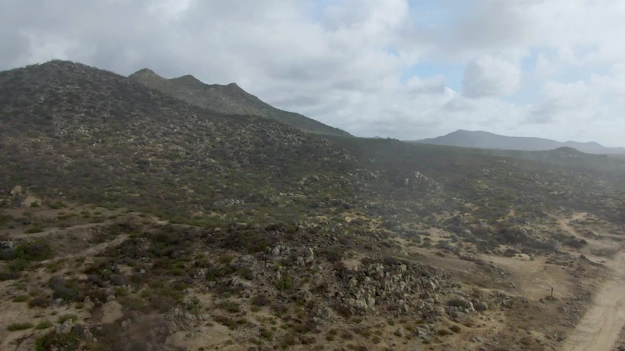 Aerial view of rocky hills and a pale dirt road in Punta Gordo, Baja California Sur, with sparse scrub, distant peaks, broken clouds, and no visible people.