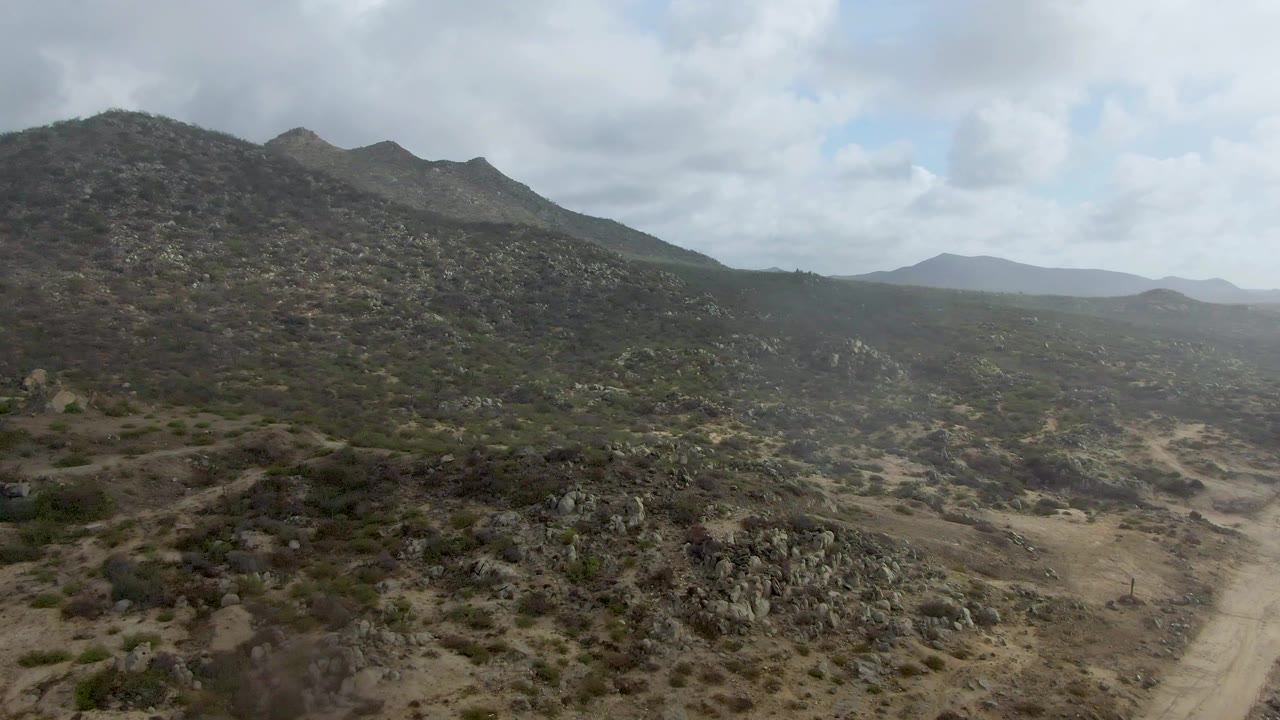 A high aerial frame over Punta Gordo terrain shows a scrub-covered mountain ahead, rocky ground below, a winding dirt road to the right, and no visible people.