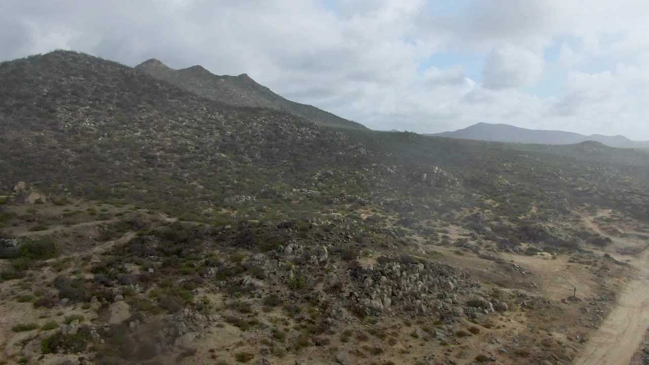 The Punta Gordo aerial view holds on a broad rocky hillside, tan dirt road, scattered shrubs, hazy distant ridges, partly cloudy sky, and no visible people.