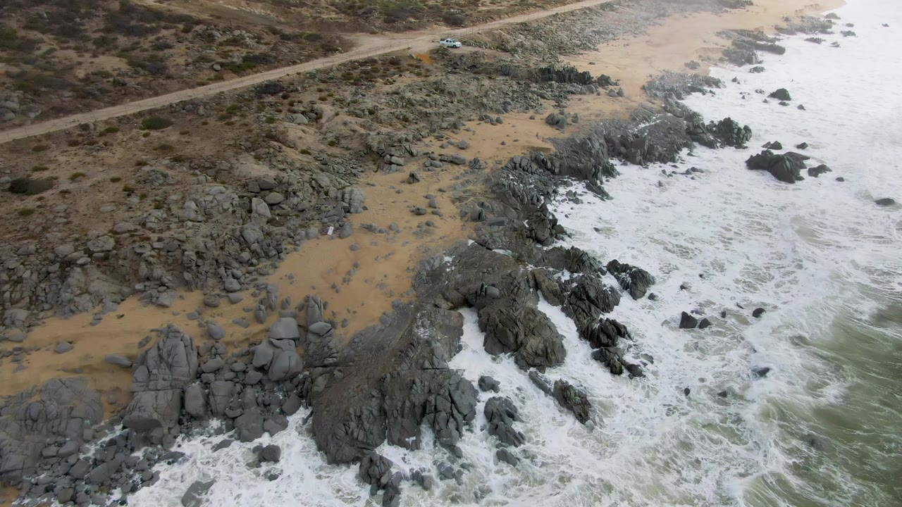 Aerial view of waves breaking against rocks beside tan sand.