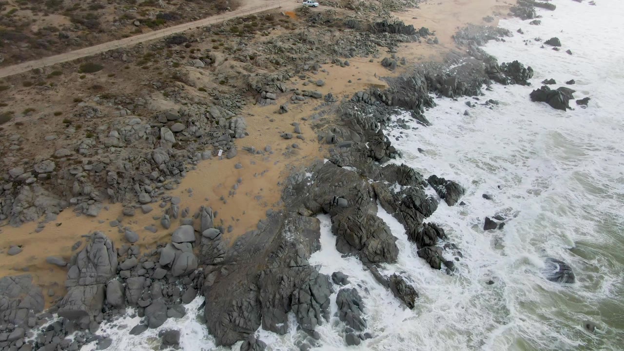 Aerial frame over Punta Gordo, Baja California Sur, with foamy water pressing into jagged rock formations and tan beach, while the inland road remains visible near the upper edge and no people are visible.