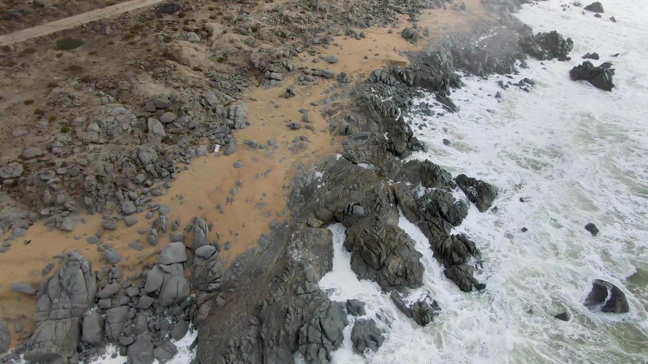 Aerial frame over Punta Gordo, Baja California Sur, showing the surf line filling more of the right side as white foam wraps around offshore rocks, with tan sand and no visible people.