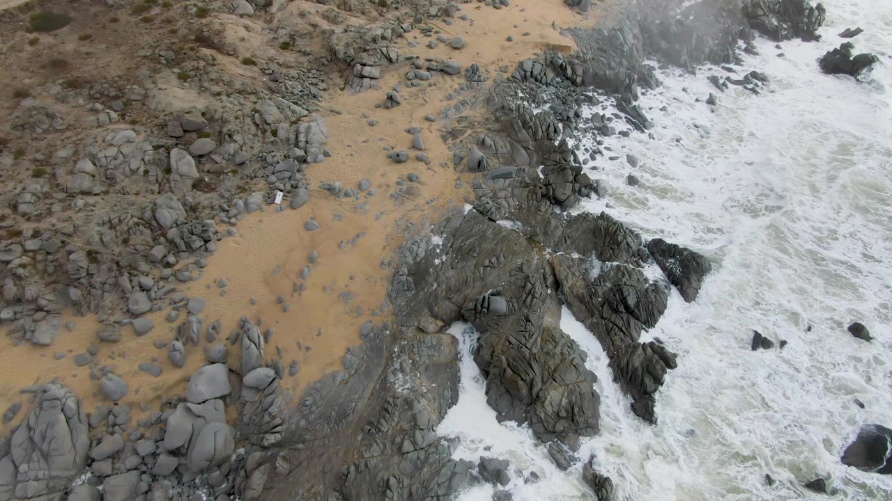 Aerial frame over Punta Gordo, Baja California Sur, where large rounded rocks and jagged outcrops fill the lower left while foamy surf covers the right side, with no visible people.