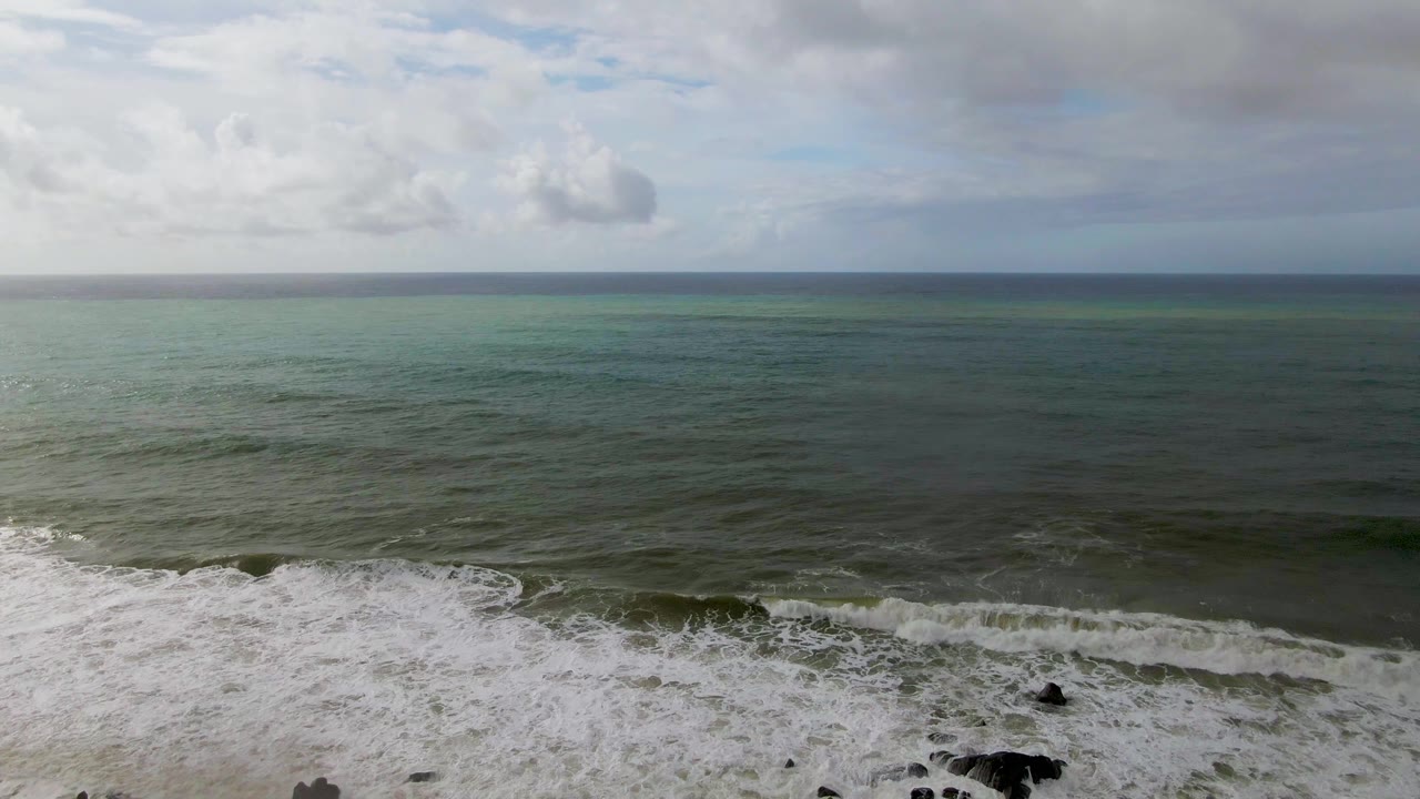 First frame of Punta Gorda, Baja California Sur surf, with white foam crossing dark rocks at the bottom, green-blue ocean beyond, cloudy sky above, and no visible people.
