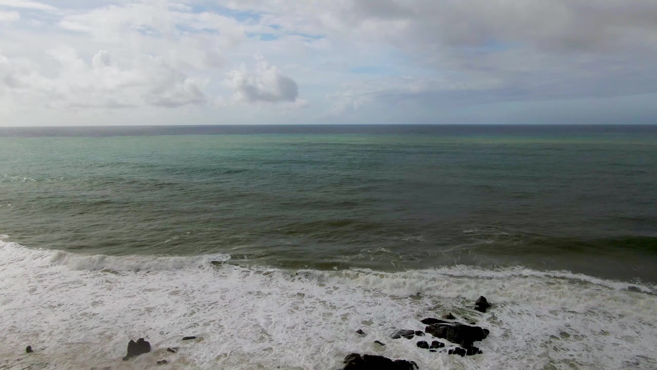 At 0:02 in Punta Gorda, Baja California Sur, dark rocks cluster near the lower right as a low wave band rolls in under a gray cloud layer, with no visible people.