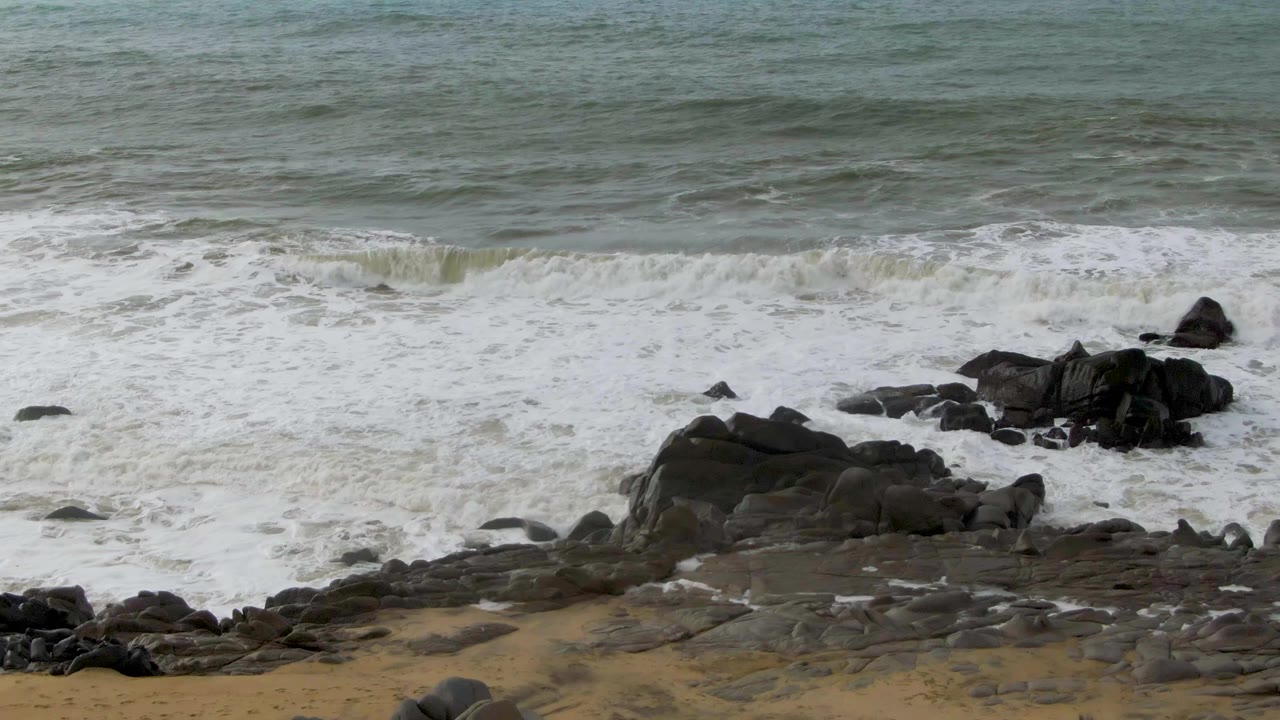 Gray-green surf breaks across dark rock outcrops and tan sand at Punta Gorda in Baja California Sur, with no visible people.