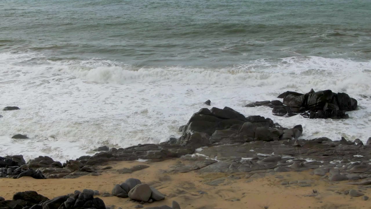 White foam spreads around black coastal rocks while wet stone ledges and sandy patches line the Punta Gorda shoreline, with no visible people.