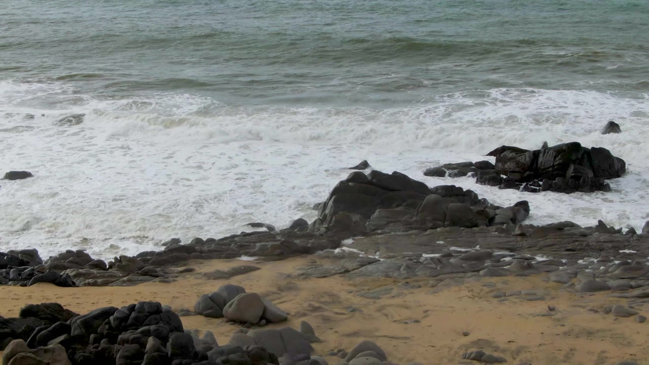 A fresh band of surf rolls toward clustered dark rocks and low sand at Punta Gorda, Baja California Sur, with no visible people.